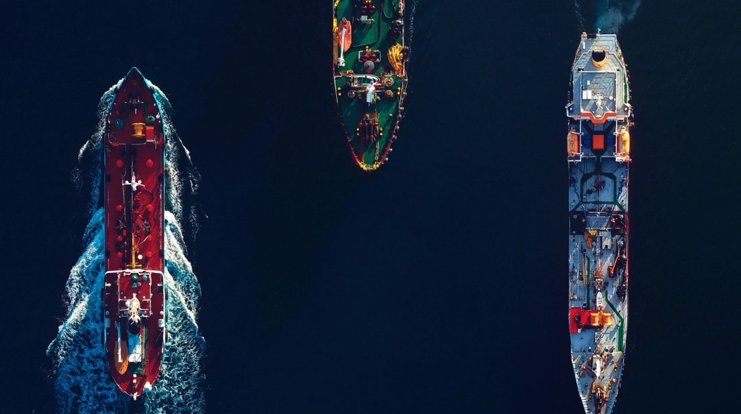 Aerial view of three cargo ships sailing on dark blue water, positioned side by side. The ship on the left is making waves, indicating movement.