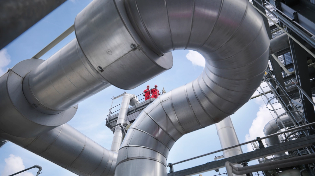 Curved metal pipes at an industrial facility with two workers in red gear standing on a platform.