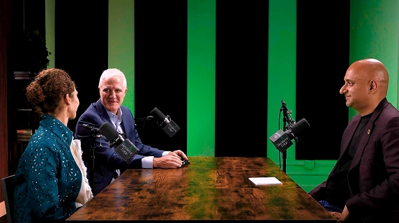 Three people engage in a podcast recording at a wooden table with professional microphones, against a green-lit background, creating a focused and lively atmosphere.