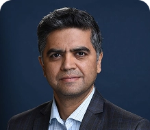 Person with short dark hair, neutral expression, wearing a dark plaid blazer over a light blue checkered shirt. Solid dark blue background.