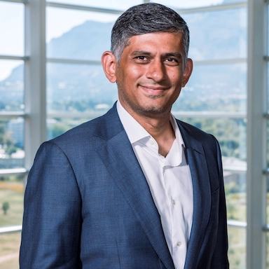 Person in a dark blue suit jacket and white dress shirt standing in front of a large window. Background shows mountains and greenery.